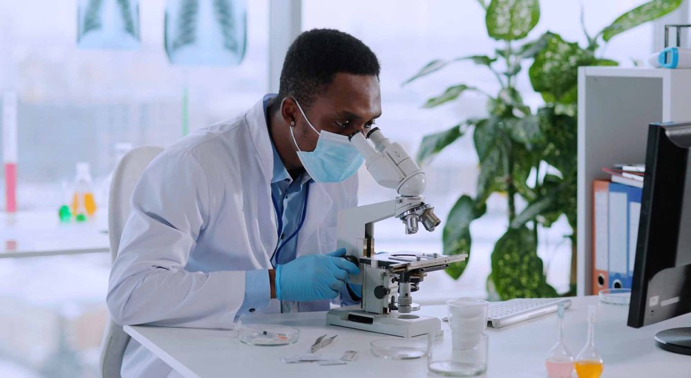 Masked researcher analyzes sample under lab microscope