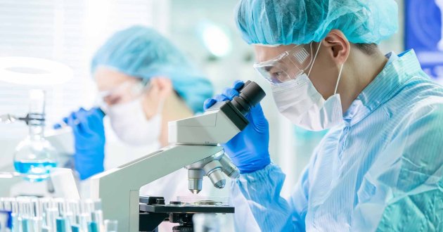 Masked researcher analyzes sample under lab microscope