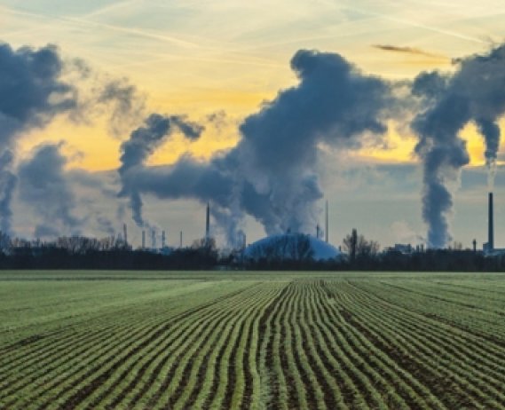 CO2 bi products entering air from smoke stacks next to a field of crops.