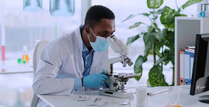 Masked researcher analyzes sample under lab microscope