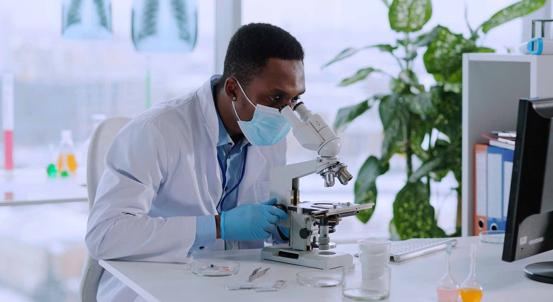 Masked researcher analyzes sample under lab microscope