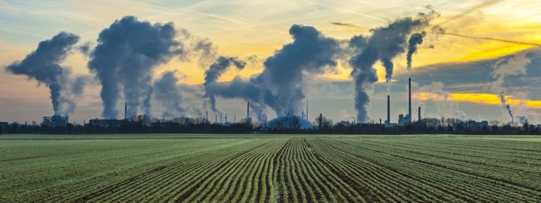 CO2 bi products entering air from smoke stacks next to a field of crops.